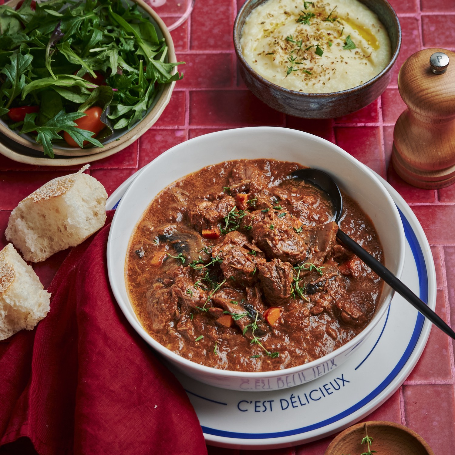 A bowl of bœuf bourguignon topped with thyme, served with crusty bread, a green salad and a bowl of celeriac purée.